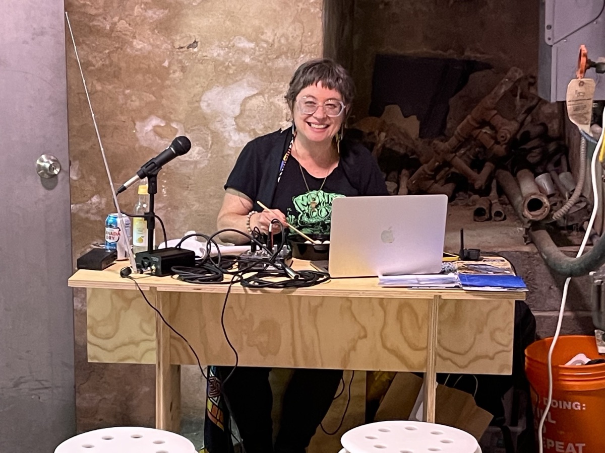 Greta Byrum smiles from behind a plywood desk with a microphone, mixer and laptop, two white stools in front, in a basement space
